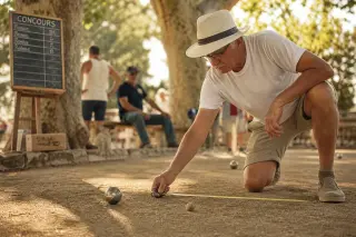 Elderly man measures distance between pétanque balls on a sunlit, shaded court with plane trees and a handwritten scoreboard in the background.